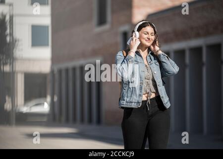 Smiling female commuter listening music through headphones while walking on street in city Stock Photo