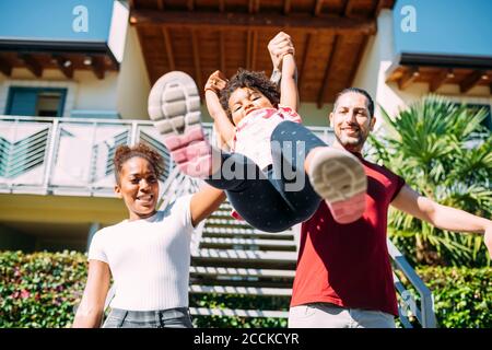 Happy parents holding playful daughter's hands and picking her up against house Stock Photo