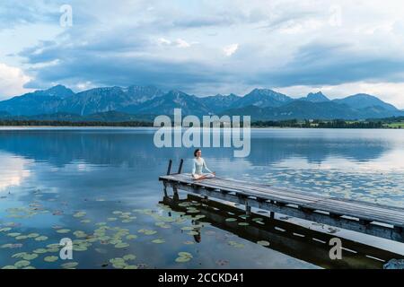 Woman meditating while sitting on jetty over lake against mountains Stock Photo