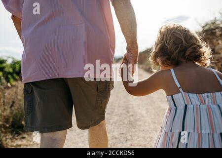 Close-up of grandfather holding granddaughter's hand while walking on dirt road