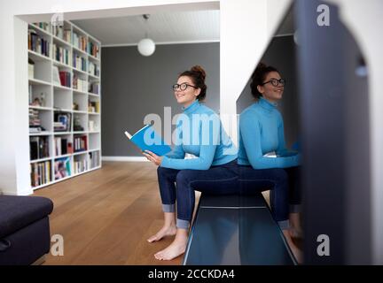 Smiling female student holding book and looking away while sitting by television set at home Stock Photo