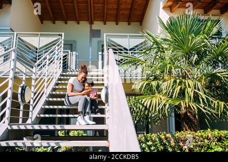Mother with daughter using smart phone while sitting on steps against house Stock Photo