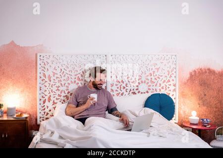 Smiling mid adult man holding coffee using laptop while relaxing on bed at home Stock Photo
