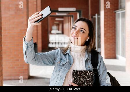 Smiling university student with book taking selfie in campus Stock Photo