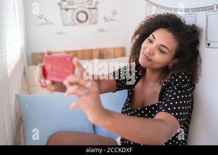 Young woman with curly hair taking selfie with smart phone while sitting against wall at home