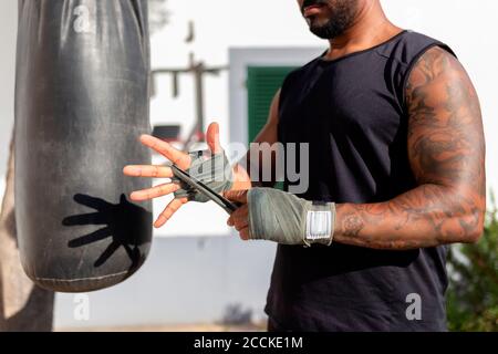 Close-up of man tying bandage on hand while standing by punching bag in yard Stock Photo