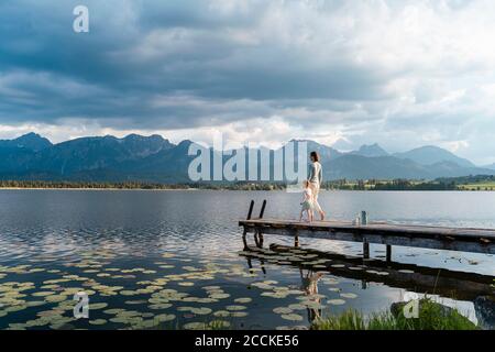 Mother and daughter walking on jetty over lake against cloudy sky Stock Photo