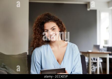 Smiling beautiful young woman with long brown curly hair holding digital tablet at home Stock Photo