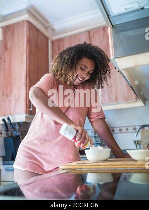 Smiling young woman with curly hair adding whipped cream in bowl at home Stock Photo