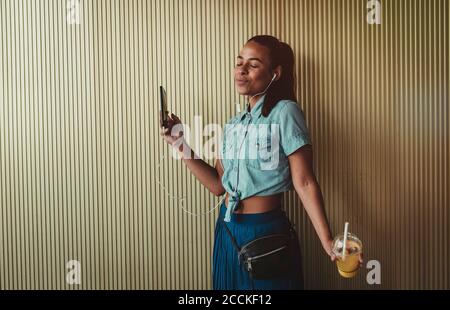 Woman with eyes closed holding drink while enjoying music through headphones against wall Stock Photo
