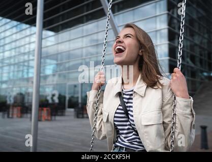 Happy young woman screaming while swinging against modern building in city Stock Photo