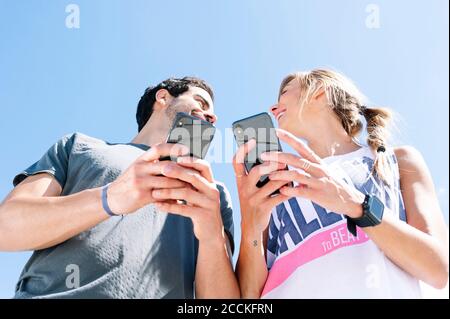 Smiling couple looking at each other while using smart phone against clear blue sky Stock Photo