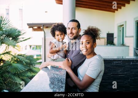 Smiling parents standing with daughter sitting on retaining wall in balcony Stock Photo