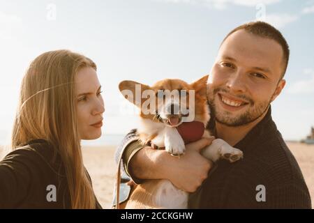 Young happy couple with dog taking selfie on beach. Beautiful woman and man and Corgi puppy are having fun. Family life, togetherness, husband and wife relax together in summer by sea Stock Photo