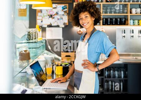 Confident female owner standing with laptop at counter in coffee shop Stock Photo