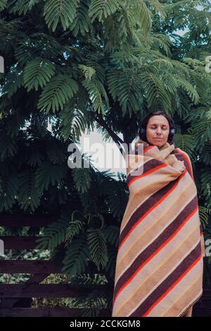 Woman with eyes closed wrapped in blanket listening music through headphones by acacia dealbata tree Stock Photo