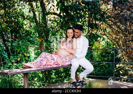 Happy man with pregnant woman relaxing on bench in park Stock Photo
