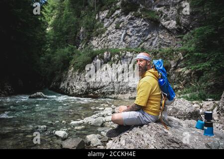 Hiker with full beard and yellow hoodie sitting on stone next to river Stock Photo
