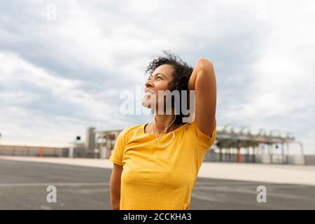 Smiling thoughtful woman looking up while standing against cloudy sky in city