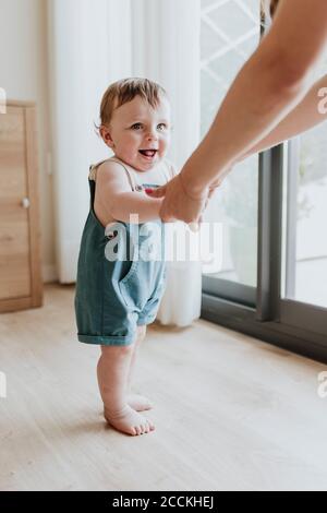 Baby girl holding mother's hands while learning to walk on floor at home Stock Photo