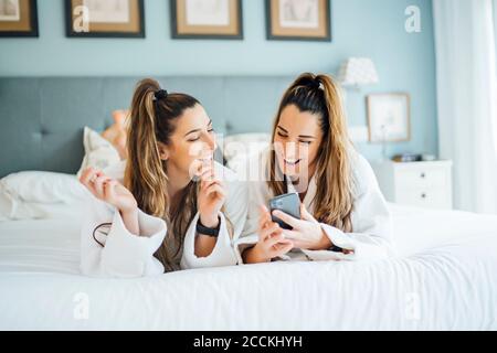 Smiling twin sisters using smart phone while lying on bed in hotel Stock Photo