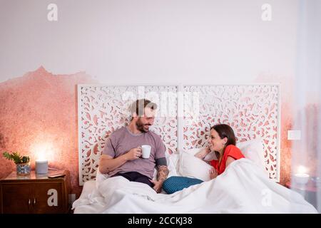 Smiling man holding coffee talking with wife while relaxing on bed at home Stock Photo