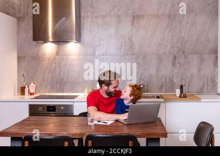 Smiling father with son using laptop on dining table in kitchen at home Stock Photo