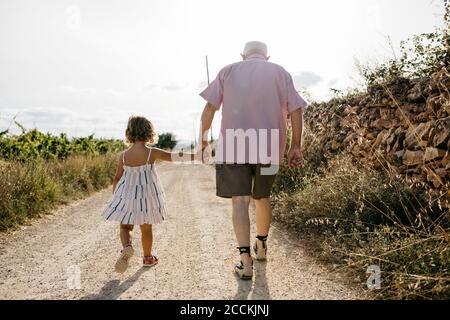 Grandfather holding granddaughter's hand while walking on dirt road against sky Stock Photo