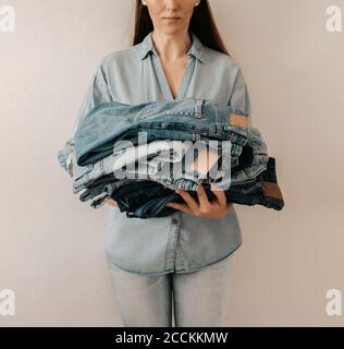 Unrecognizable woman holds stack of lot jeans pants on white background. Caucasian woman with long brown hair weared in blue shirt and jeans holds heap of denim pants with differents shades of blue Stock Photo