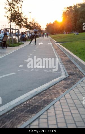 A new bike path with asphalt and a girl rolling along it in the evening on the embankment next to people resting in the rays of the setting sun.