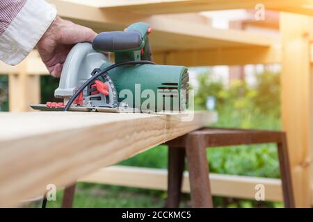 A working electric saw of green color leading with a hand along a wooden beam during construction at a summer cottage on a summer day. Stock Photo