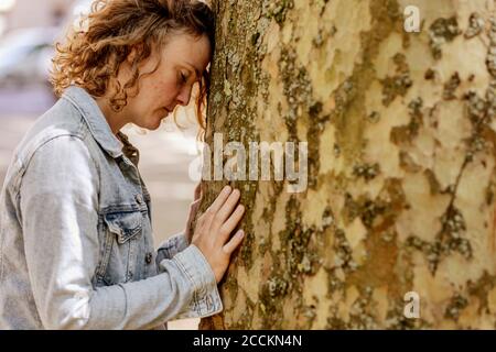 Sad mid adult woman with eyes closed leaning on tree trunk in park