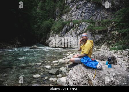 Hiker with full beard and yellow hoodie sitting on stone next to river and using smartphone Stock Photo