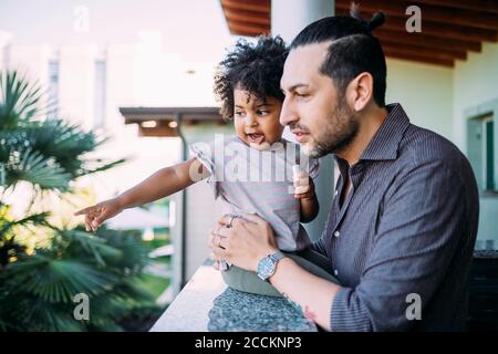 Father holding baby girl pointing on retaining wall while standing in balcony Stock Photo