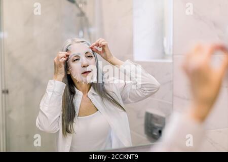 Portrait of beautiful gray haired retired woman in bathroom, removing white sheet mask from her face, getting home antiwrinkle moisturizing beauty Stock Photo