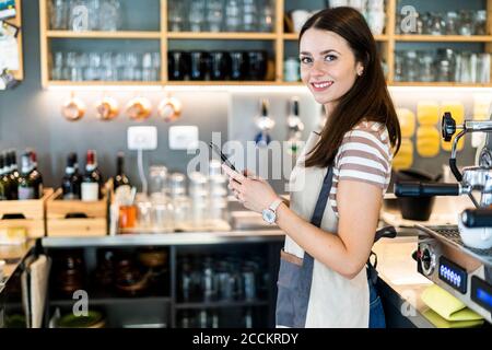 Happy female owner holding smart phone while standing in coffee shop Stock Photo