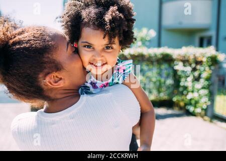 Mother kissing happy daughter while carrying her on footpath during sunny day Stock Photo