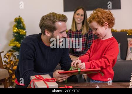 Smiling woman looking at father giving Christmas present to son in living room Stock Photo