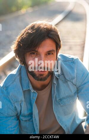 Close-up of handsome man wearing denim jacket sitting against railroad track in city Stock Photo