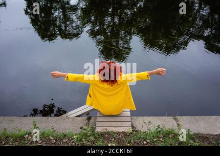 Young woman with curly hair and yellow suit sitting by the riverside with arms outstretched Stock Photo