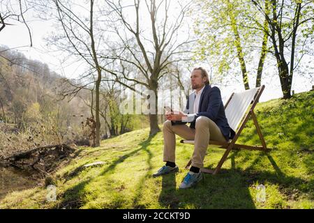 Thoughtful businessman holding smart phone while sitting on chair against trees at park Stock Photo