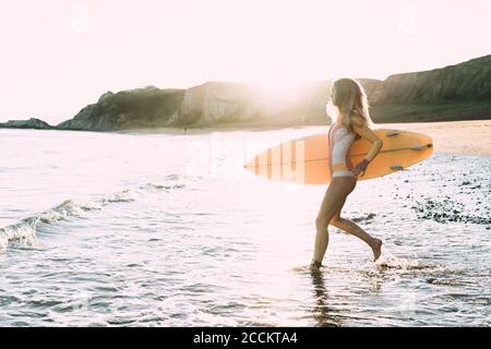 Young woman with surfboard running in sea against clear sky during sunset Stock Photo