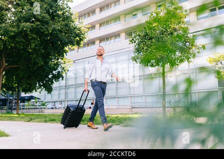 Businessman walking with wheeled luggage at office park Stock Photo