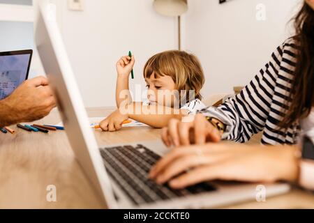 Cute daughter painting on paper while sitting with working parents in dining room Stock Photo