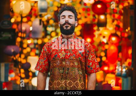 Attractive bearded man with colorful shirt standing in front of lamps with eyes closed Stock Photo