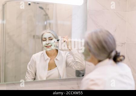 Pretty senior lady in white shirt smearing beauty clay mud mask on her face, while standing in front of mirror during spa procedure at home. Skin care Stock Photo