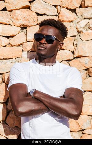 Close-up of young man wearing sunglasses with arms crossed standing against stone wall Stock Photo