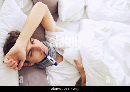 Sick young man with thermometer in mouth resting on bed at home Stock Photo