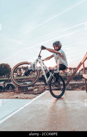 Young man wearing helmet riding bicycle on ramp in park against sky during sunset Stock Photo