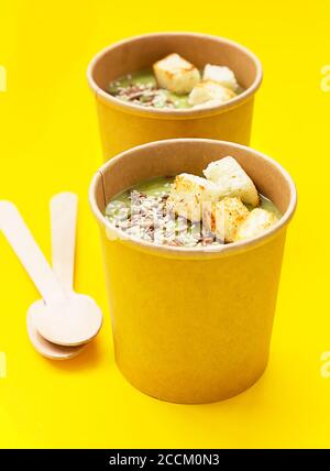 Fresh broccoli cream soup with croutons and seeds in craft containers on yellow background. Soup to go, healthy food delivery. Stock Photo